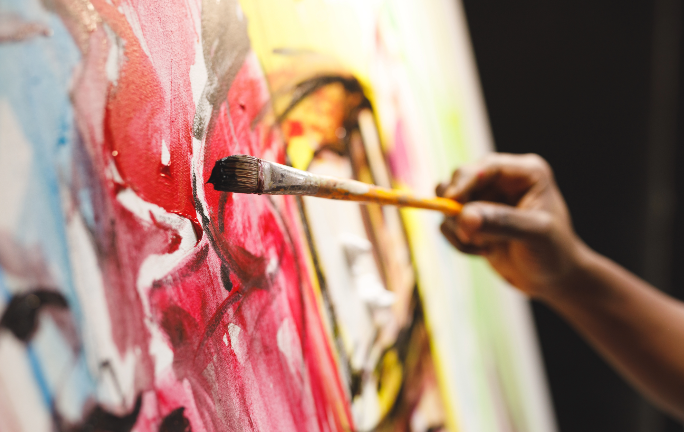 A hand belonging to a male person with brown skin paints in bold, abstract strokes on a canvas.